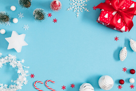 Christmas and New Year frame background with red gift box, star candle, mini trees, and ornaments on light blue backdrop. Festive winter holiday border flat lay with central copy space for text.