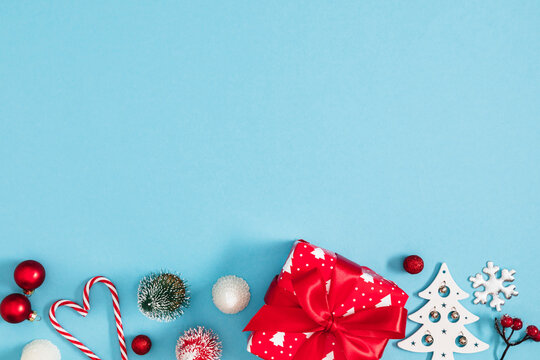 Christmas and New Year background with red gift box, wooden tree, and candy canes on light blue backdrop. Festive winter holiday bottom border composition with large copy space for text, top view.