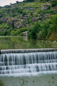 View of Shere Hills featuring a cascading waterfall over a stone weir, surrounded by lush green vegetation and rocky hills in Shere Hills, Plateau, Nigeria.