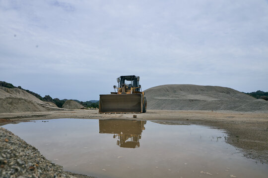 View of a yellow bulldozer parked in a quarry with piles of gravel and a reflective puddle in the foreground under an overcast sky in Shere Hills, Plateau, Nigeria.