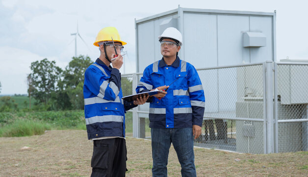 Wind energy technician reports by handheld radio while an engineer holds inspection documents, coordinating field observations beside utility equipment during a renewable power site check.