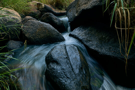 View of a small stream flowing over dark volcanic rocks in the Kerang region, with lush green grasses along the banks in Kerang, Plateau, Nigeria.
