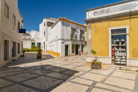 View of a sunny street with traditional cobblestone pavement, white-washed buildings with yellow accents, and a shop window displaying ceramics in Tavira, Faro District, Portugal.