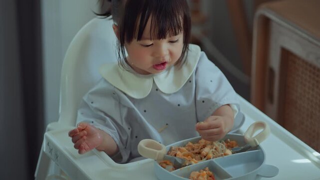 Cute Asian toddler girl eating independently while sitting in high chair at home, little child using fingers during mealtime with food tray, self feeding.