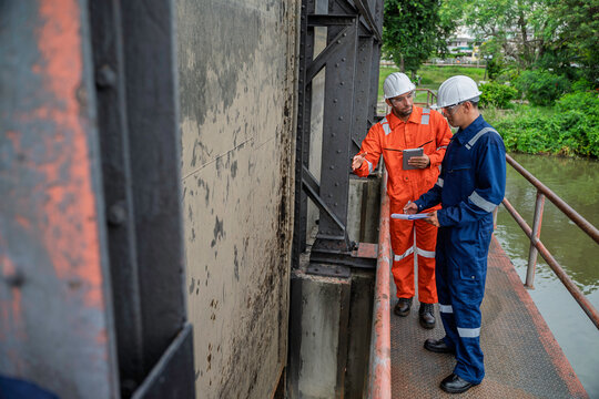 Industrial engineer and technician inspect a floodgate structure at a water control facility, reviewing documents and discussing gate condition, flood control, water management, and repair work.