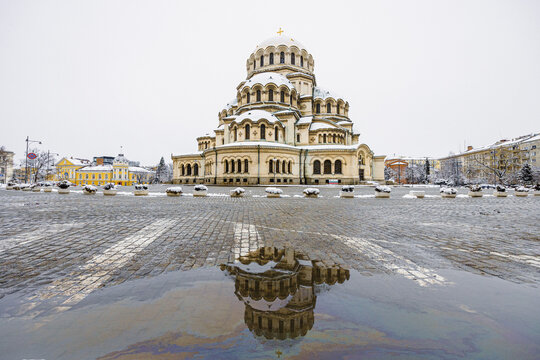 View of Alexander Nevsky Cathedral with snow-covered ground and a reflection in a puddle on the cobblestone square in Sofia, Sofia City Province, Bulgaria.