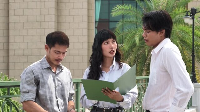 Three young Asian business colleagues discussing a project while looking at a green document folder outdoors in front of a building.