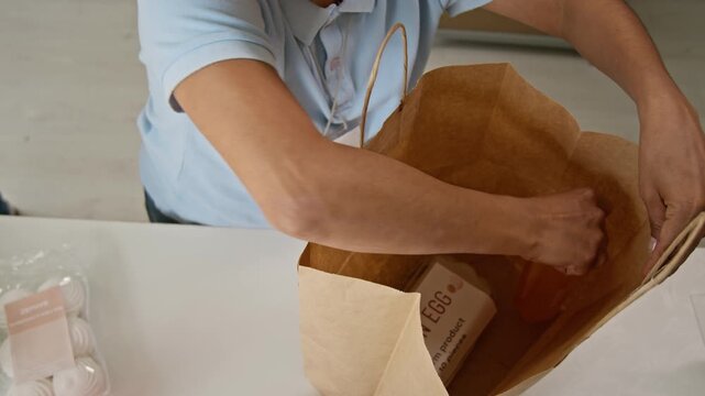High angle shot of unrecognizable female worker packing eggs, bottled drinks and organic groceries into paper bag. Shot ideal for online supermarket delivery and retail advertising