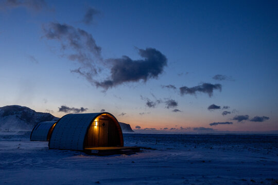 View of barrel-shaped cabins illuminated at dusk in a snowy landscape in Hvolsvollur, Rangarthing eystra, Iceland.