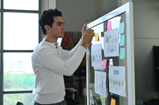 A young man focusing on a whiteboard filled with colorful post-it notes and graphs, brainstorming ideas in a modern office environment.