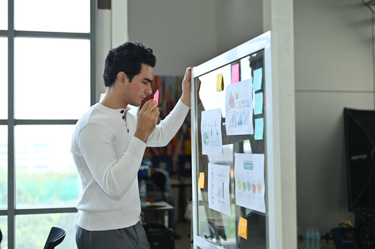 A young man thoughtfully analyzes a project on a whiteboard filled with colorful sticky notes, demonstrating creative problem-solving and teamwork.