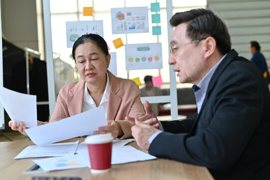 Two professionals collaborate on a project, reviewing documents and discussing ideas in a modern office environment with charts in the background.