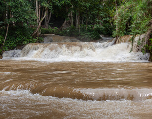 Kuang Si Waterfall flows with muddy brown water over limestone cascades surrounded by dense tropical forest. © Eugene Ga