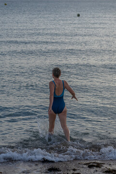 Langrune-Sur-Mer, France - 08 09 2026: A mature woman wearing a blue swimsuit from behind walks in the water to get in the sea
