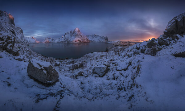 View of the snow-covered peaks of the Lofoten mountains reflected in the calm waters of the fjord at twilight in Norway.