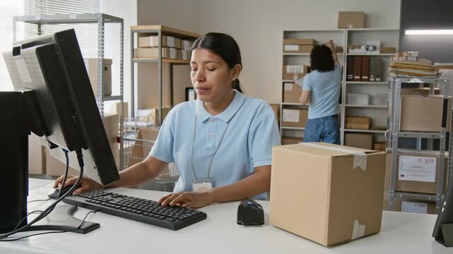 Hispanic young woman scanning parcel barcode with handheld scanner and typing data into computer at logistics center. Ideal for supply chain, ecommerce, and order fulfillment campaigns
