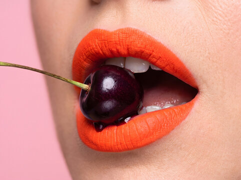 Woman with bold red lipstick biting dark cherry on green stem against pink background