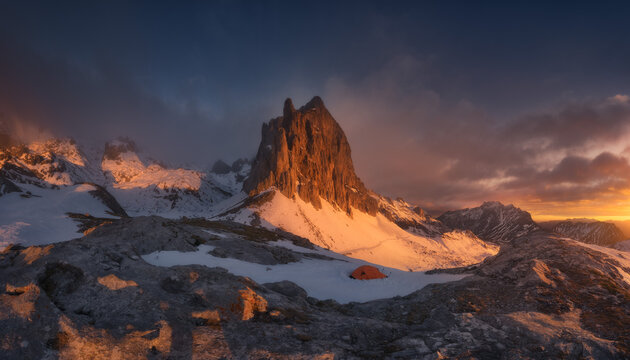 View of a small orange tent pitched on a snow-covered mountain ridge during golden hour with dramatic peaks and clouds in the background in Spain.