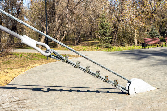 Steel suspension cable anchored with a thimble and a row of U-bolt clamps descending to a rounded concrete pylon on a pedestrian bridge deck in a sunny riverside park with a wooden bench nearby.