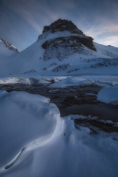 View of a snow-covered mountain peak rising above a frozen stream with icy textures under a soft twilight sky in Spain.