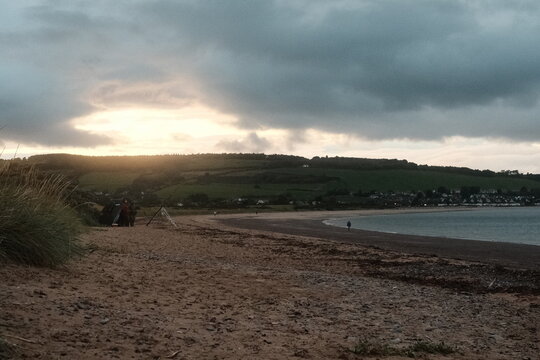 View of Chanonry Lighthouse on the coast with sandy beach, grassy dunes, and rolling hills under a dramatic cloudy sky at sunset in Fortrose, Scotland, United Kingdom.
