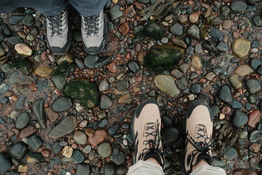 View of two pairs of hiking boots standing on a rocky beach with smooth pebbles and patches of seaweed in Fortrose, Scotland, United Kingdom.