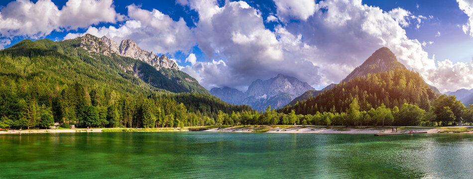 View of Lake Jasna with turquoise water, surrounded by lush green forests and the towering Julian Alps under a bright sky with fluffy clouds in Kranjska Gora, Jesenice, Slovenia.
