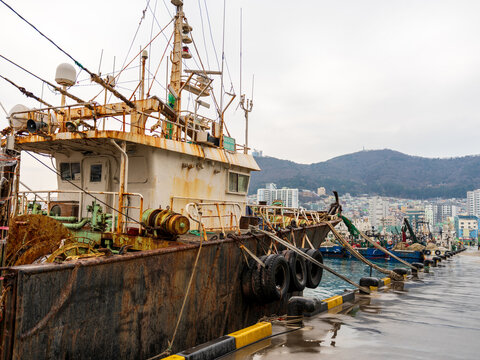 View of a rusty fishing boat docked at the harbor with tires as fenders, calm water, and a cityscape with hills in the background in Busan, Busan, South Korea.