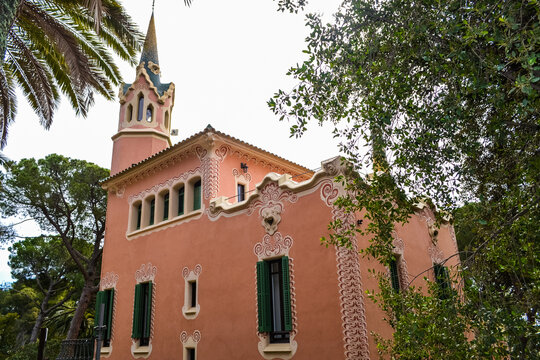 View of Casa del Guarda in Park Guell with its pink facade, ornate architectural details, green shutters, and a tall turret, set amidst trees in Barcelona, Catalonia, Spain.