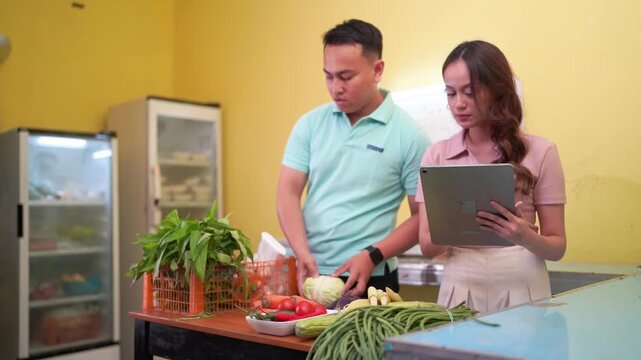 Light blue polo man reaches across produce table while pink top woman checks tablet, indicating vegetable sorting in bright market stall with warm yellow walls and glass refrigerator cases.