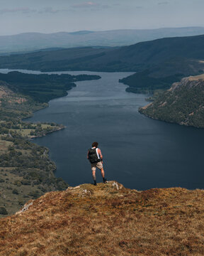 View of a hiker with a backpack standing on the rocky summit of Sheffield Pike overlooking the expansive Ullswater lake surrounded by rolling hills in Penrith, England, United Kingdom.