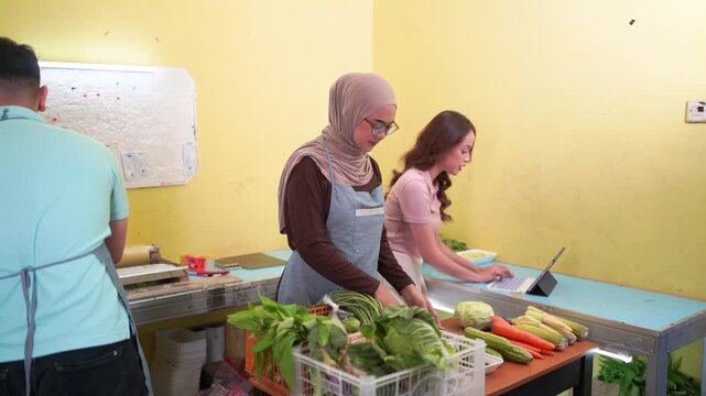 Three young adults sort fresh vegetables in simple indoor produce workspace, lifting cabbage and leafy greens into baskets while one checks tablet beside trays.