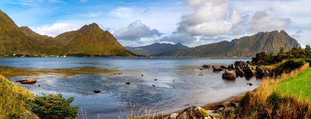 Wide panorama near Sennesvik in Lofoten with calm blue sea, rocky shore, green vegetation and impressive mountains beneath a partly cloudy blue sky. © EKH-Pictures