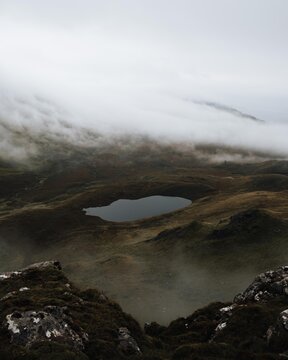 View of The Storr landscape featuring a small loch nestled among rugged hills under a blanket of low-hanging clouds and mist in Portree, Scotland, United Kingdom.