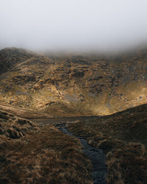View of Eryri National Park with rocky mountain slopes, a small stream, and misty clouds in Wales, United Kingdom.