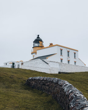 View of Stoer Lighthouse with its white walls and dark lantern room set against a grassy hill and a stone wall in the foreground in Lairg, Scotland, United Kingdom.