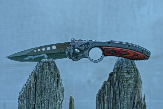 A folding sharp knife with a metal blade and a wooden handle rests on two wooden supports. Old model with noticeable signs of wear and scratches on the surface.