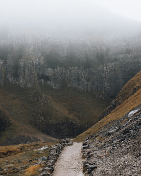 View of Gordale Scar featuring a narrow stone-lined path leading towards towering limestone cliffs shrouded in thick mist and autumn grass in Malham, England, United Kingdom.