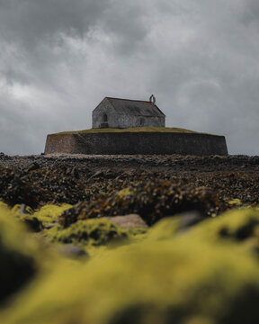 View of St Cwyfan's Church surrounded by stone walls on a tidal island under a moody overcast sky in Ty Croes, Wales, United Kingdom.