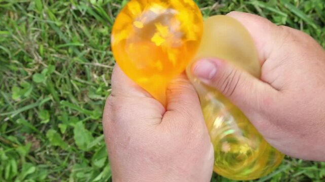 A close up shows orange and yellow water balloons being squeezed by hand on green grass, steadily deforming and stretching as fingers press, twist and reshape them outdoors in daylight.