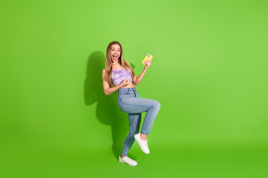 Joyful young woman jumping with smartphone in bright purple top and jeans against a vivid green background for lifestyle fashion and shopping themed stock image