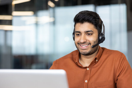 Customer support representative working on laptop, smiling man with headset providing remote assistance in office, professional communication and digital service concept for business operations.