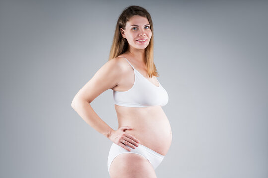 Happy pregnant woman with big belly in white underwear on gray background, pregnancy concept