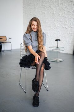Smiling young woman seated on wire metal chair in bright studio, wearing light gray wool coat over black lace dress, sheer tights, and black shoes