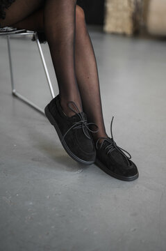 Crossed legs in sheer black stockings paired with black lace up shoes, resting on smooth studio floor under soft lighting for fashion pose and modern editorial vibe