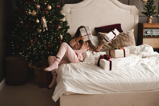 Funny child girl 5-6 year old holding Christmas present boxes in bed over xmas tree in bedroom. Winter holiday season. Selective focus.