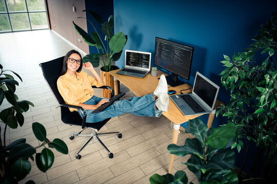 Happy female programmer working relaxed in a plant-filled home office with feet on desk, coding across multiple laptops and monitors for remote work and casual tech lifestyle