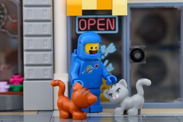 Fototapeta premium Tambov, Russian Federation - September 19, 2026 A blue Lego astronaut minifigure standing outside a shop with an Open sign, petting a grey cat while an orange cat standing nearby.