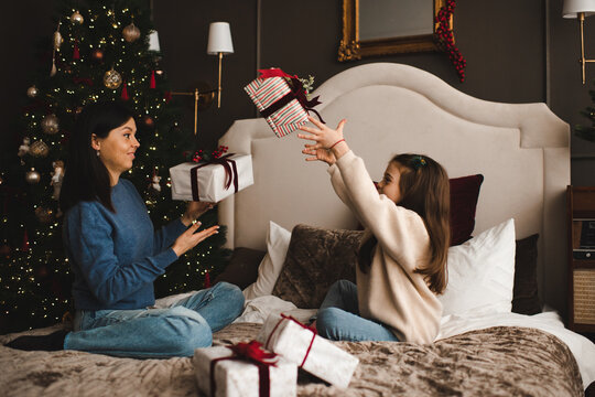 Happy woman sitting in bed with kid girl open Christmas present boxes in bedroom. Winter holiday season. Cozy home atmosphere.