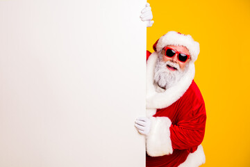 Obraz premium Santa Claus wearing red sunglasses peeking from behind a blank white sign on a yellow background — cheerful festive Christmas advertising and holiday greeting concept